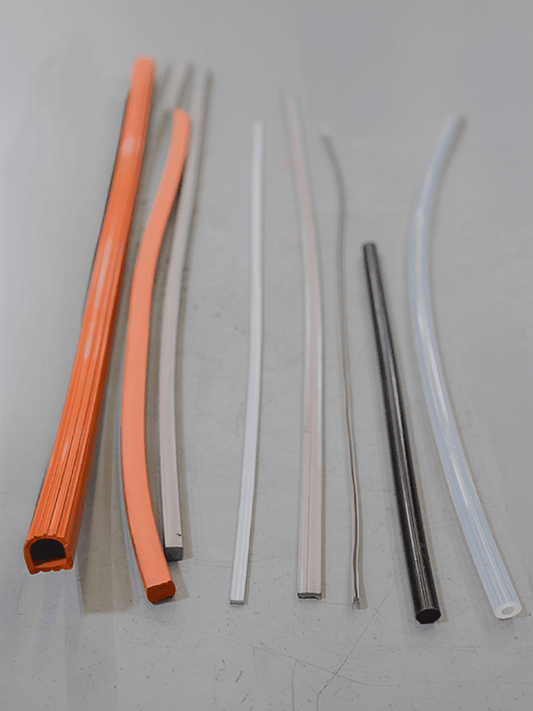 Six different colored flexible silicone hoses and rods arranged vertically on a light gray surface. The colors include orange, peach, silver, light gray, black, and white.