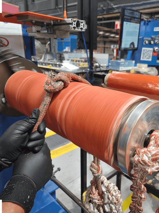 A close-up of a large orange industrial hose being handled by a person wearing black gloves in a factory setting. A thick braided rope is wrapped around the roller, and other machinery parts are visible in the background.