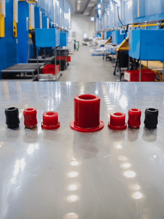 Six cylindrical red and black polyurethane bushings are arranged on a reflective metal table in a factory setting, with a blurred industrial background.
